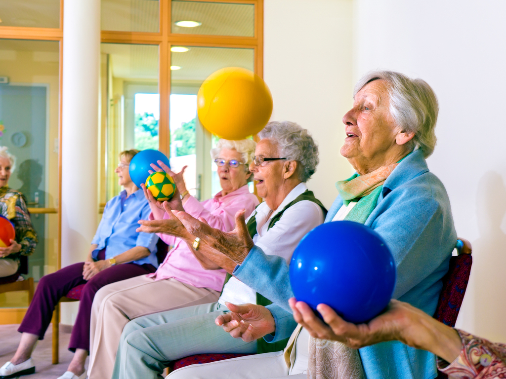 Group of happy senior ladies doing coordination exercises in a seniors gym sitting in chairs throwing and catching brightly coluored balls